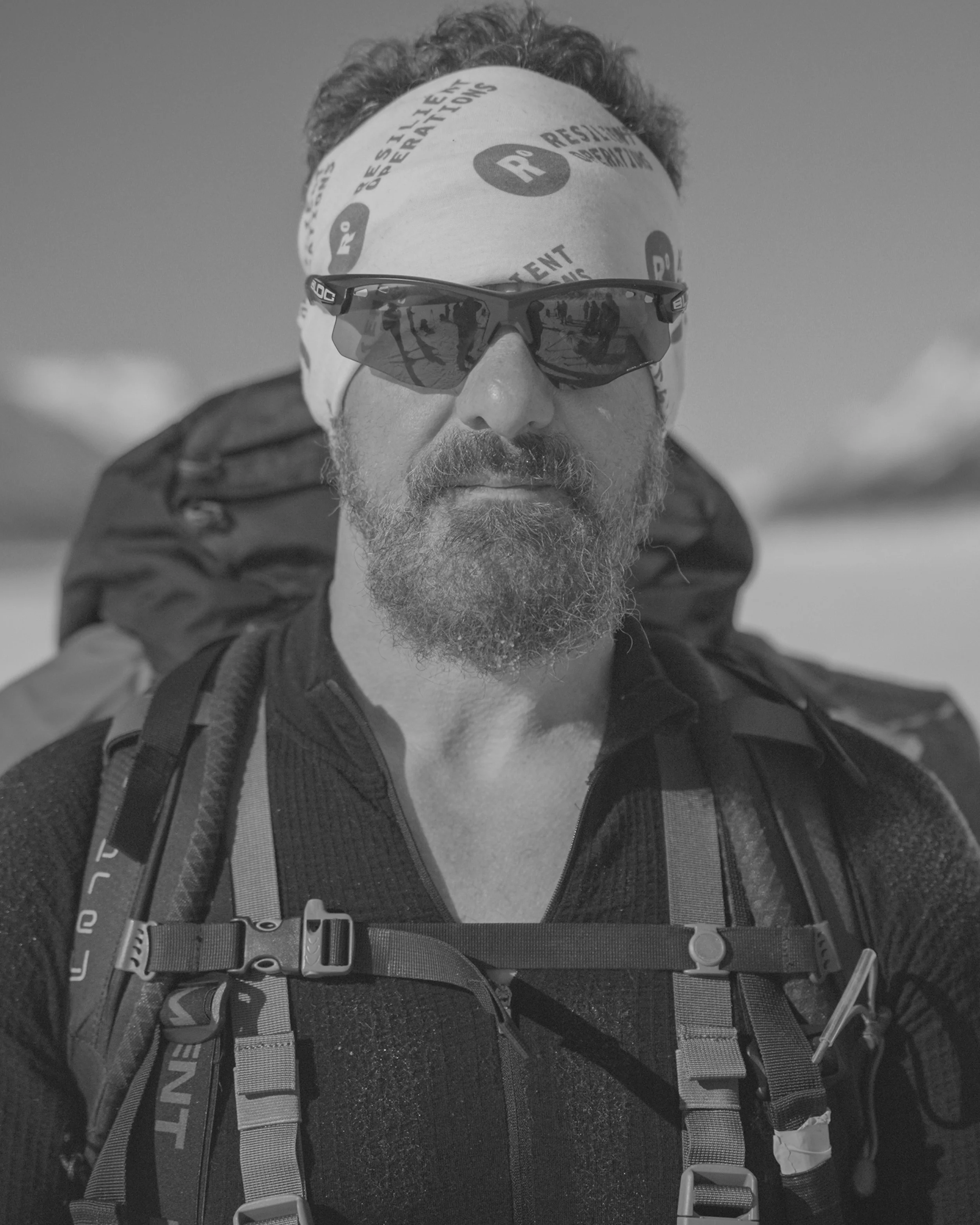 Man with curly hair and beard wearing sunglasses, a bandana, and a backpack in an outdoor setting.