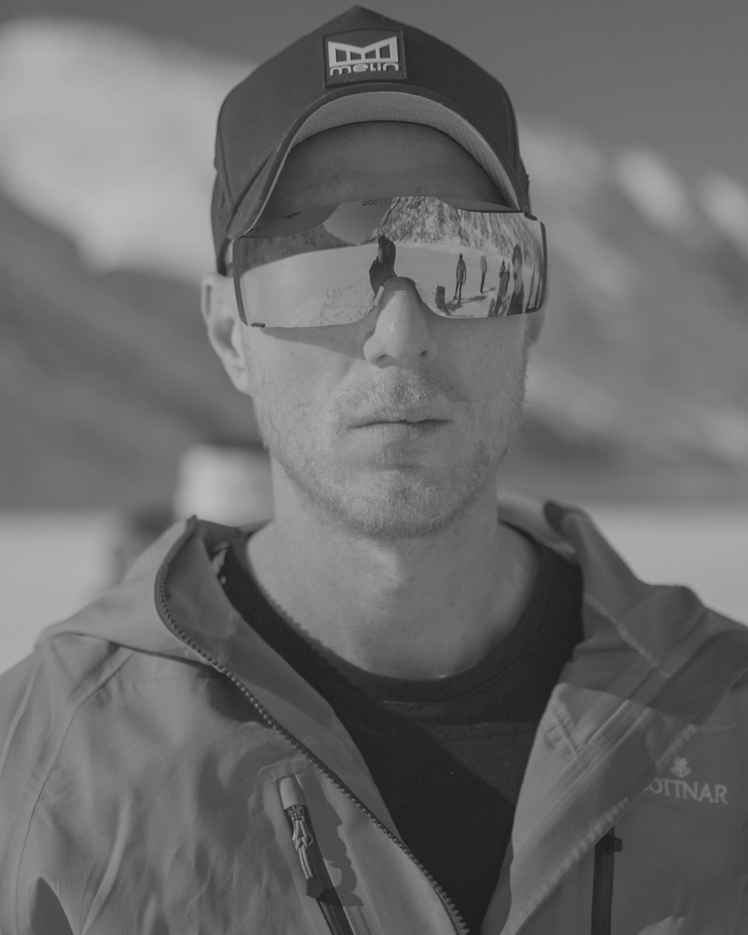 Man wearing a baseball cap, reflective sunglasses, and a jacket, with a desert landscape and other people reflected in the sunglasses.
