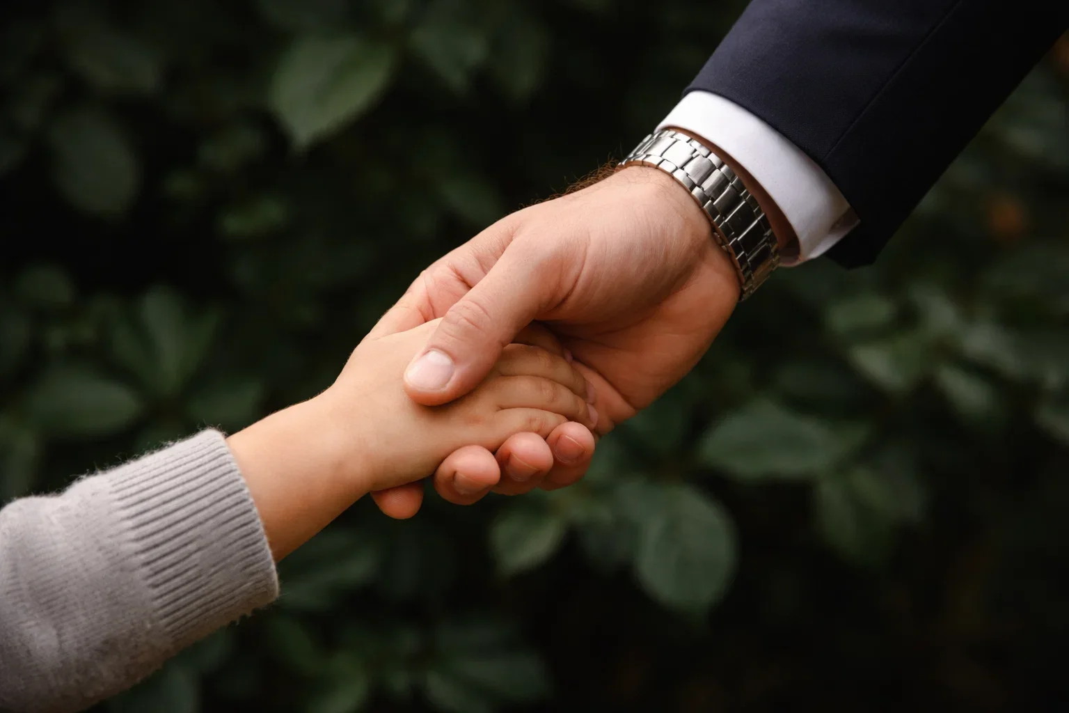 Dos manos, una adulta con reloj, la otra infantil, estrechándose en señal de ayuda o protección, con fondo de hojas verdes.