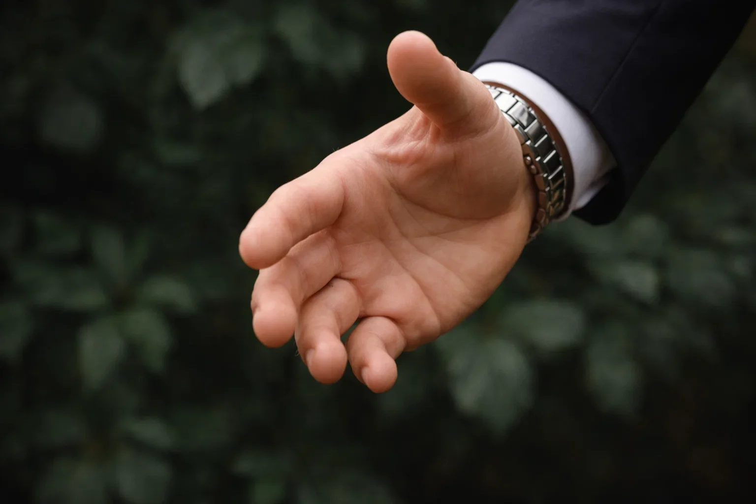 Mano extendida con la palma hacia arriba, vestido con saco y reloj en la muñeca, fondo borroso de hojas verdes.