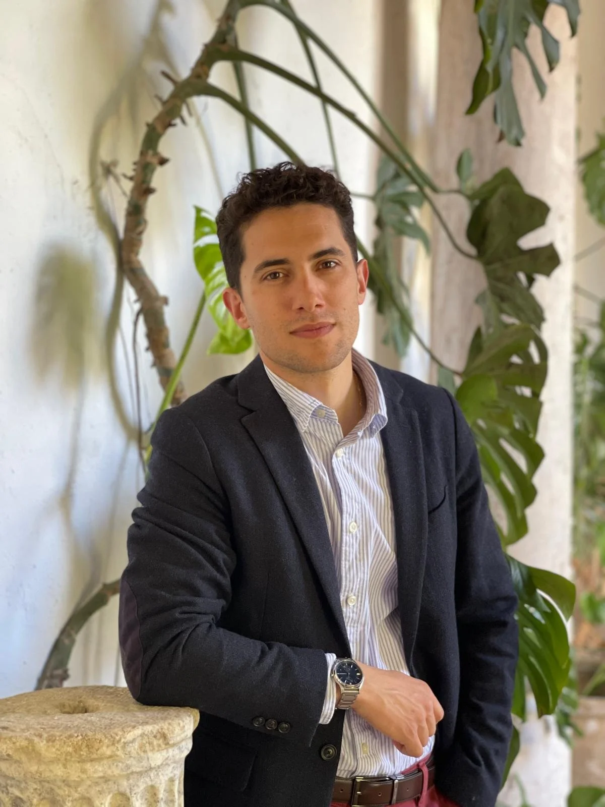 Joven hombre con cabello rizado y piel clara, vestido con saco oscuro y camisa blanca con rayas, posando con brazos cruzados al lado de plantas verdes, en un ambiente interior luminoso.