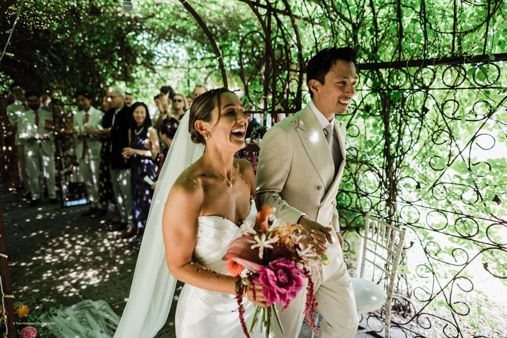Bride and groom smiling and holding hands during their wedding ceremony in a lush green outdoor setting, with wedding guests in the background.