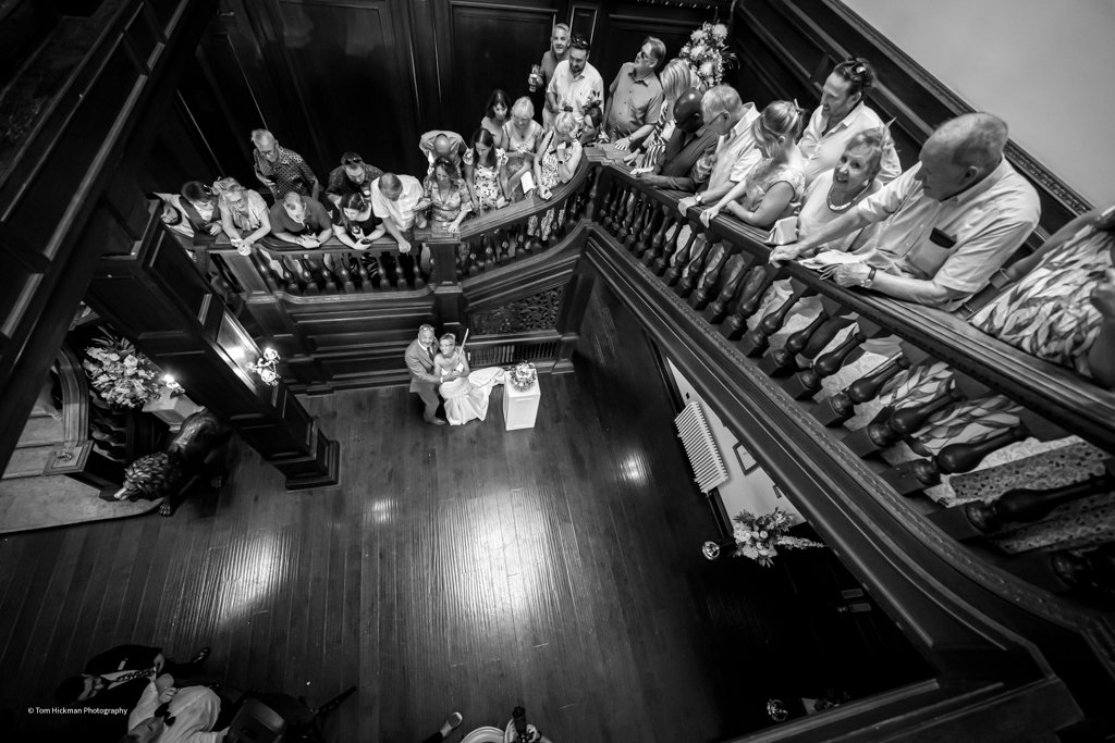 Guests gathered on an upper balcony, watching a couple at the bottom of a staircase during a wedding or formal event.