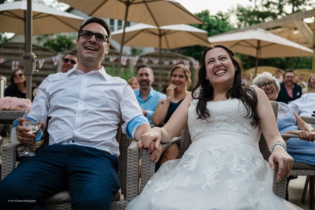 A group of people sitting outdoors under large umbrellas, smiling and enjoying a celebration, including a couple holding hands and wearing wedding attire.