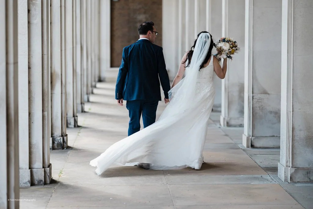 Bride and groom holding hands walking through a colonnade with stone pillars, the bride wearing a white wedding dress and veil, carrying a bouquet, with the groom in a dark blue suit.