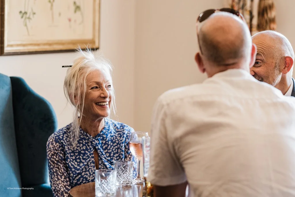 An elderly woman with white hair and a blue patterned blouse smiling and enjoying a drink, with three men engaged in conversation around her in a cozy indoor setting.