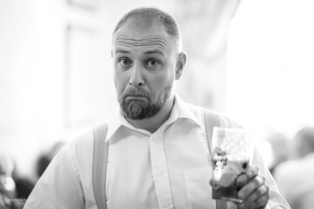 A man with a beard and short hair, wearing a white dress shirt and suspenders, holding a glass of drink, looking surprised or confused.