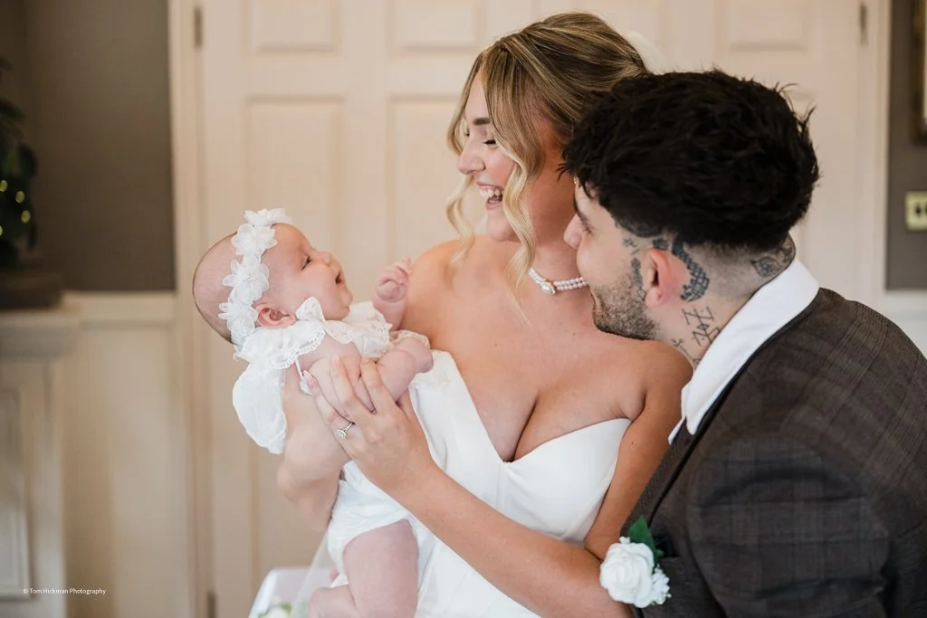 A woman in a white dress holding a baby girl dressed in white with a floral headband. The woman and a man with tattoos on his neck are smiling and looking at the baby, indoors with light-colored walls.