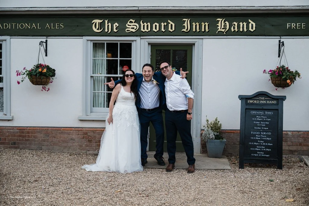 Three people, a bride in a white wedding dress and two men in suits, standing outside The Sword Inn Hand pub, smiling and posing together in front of the pub's entrance with hanging flower baskets on either side.