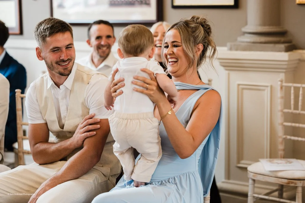 A woman in a blue dress holding a baby and smiling at him during a joyful gathering with people in the background.