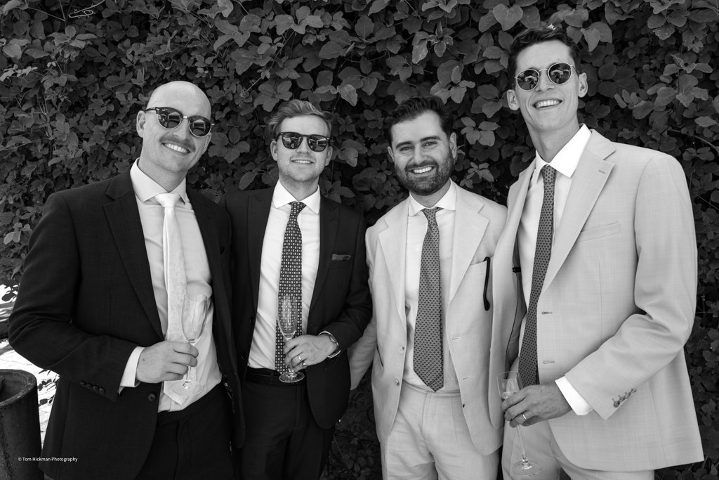 Four men dressed in suits and sunglasses smiling at the camera, holding glasses, standing in front of leafy bushes.
