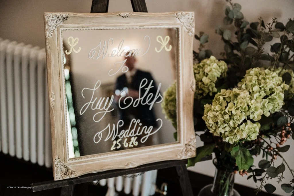 A decorative mirror with wedding welcome message written on it, placed on an easel beside a bouquet of green hydrangeas and eucalyptus leaves.