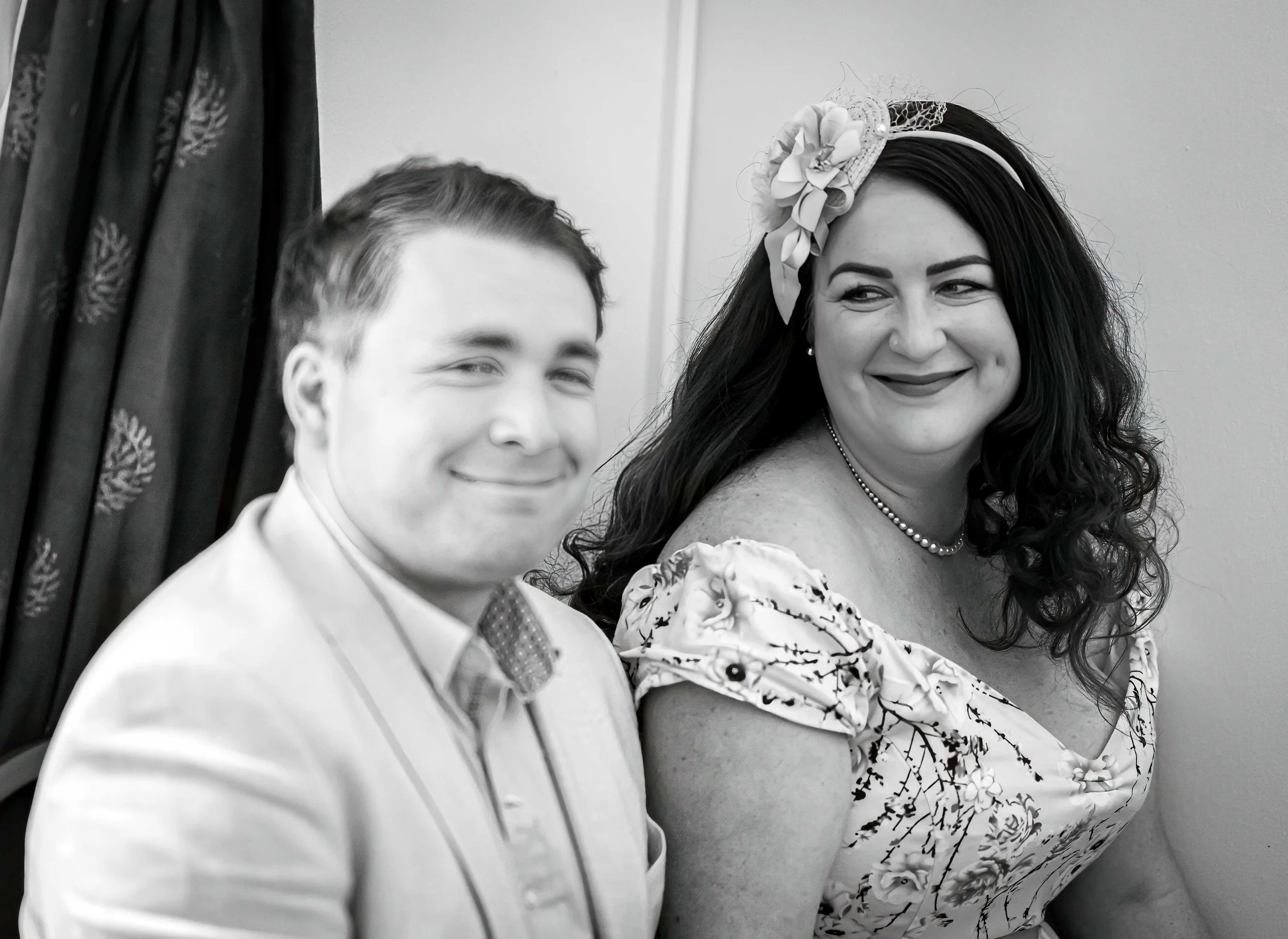 Black and white photo of two people dressed in fancy attire, smiling, with one person wearing a floral dress and a headband with a large flower and lace detail, and the other in a suit with a bow tie.