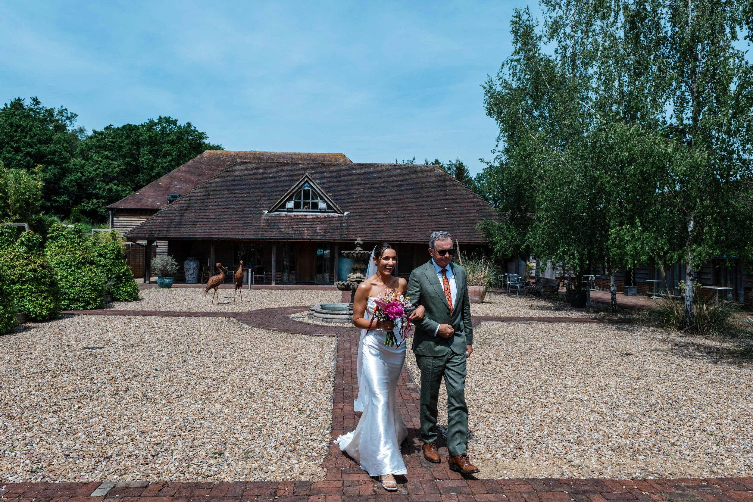 A bride and an older man, possibly her father, walking outdoors on a sunny day. The bride is wearing a white wedding gown and holding a bouquet of flowers, while the man is dressed in a suit and sunglasses. They are on a gravel pathway with greenery 