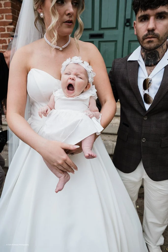 A woman in a wedding dress holding a yawning baby dressed in white, with a man standing next to them outside in front of green doors.