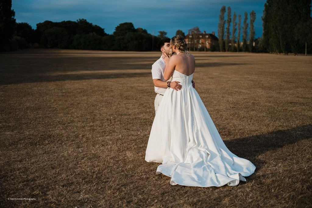 A bride and groom share a kiss in an open field at sunset, with the bride wearing a white strapless wedding gown and the groom in a white shirt and trousers.