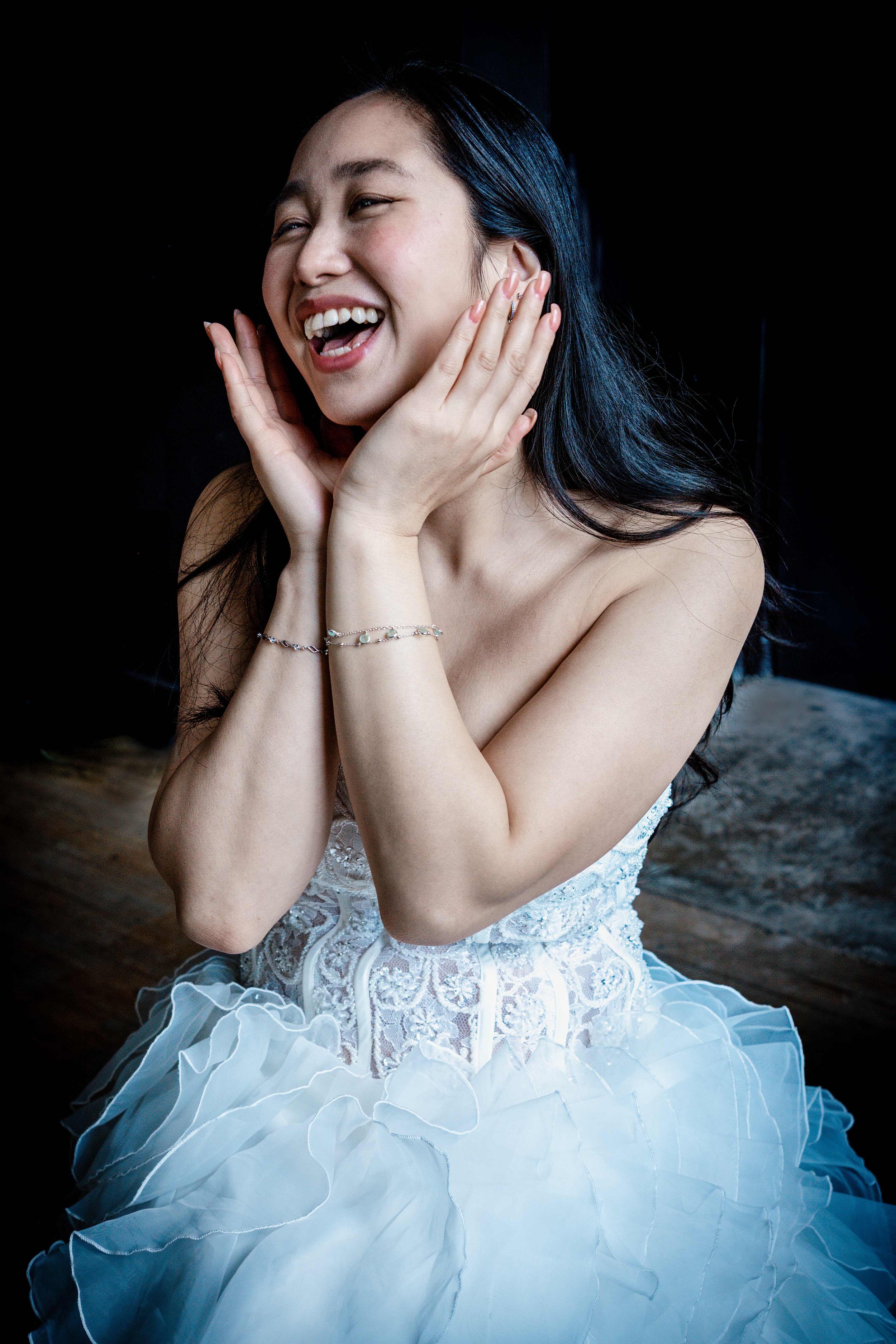 A woman with long dark hair and a broad smile, wearing a white dress with lace details and a ruffled skirt, extends her hands to her cheeks with jewelry on her wrists, against a dark background.