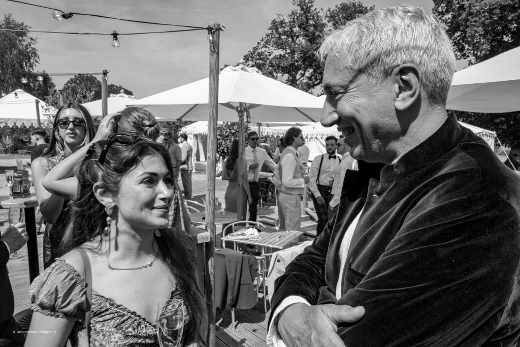 A woman and a man are smiling and engaged in conversation at an outdoor event. The woman has long hair and is wearing earrings, while the man has short light-colored hair and is wearing a dark jacket. In the background, there are other people, tents,
