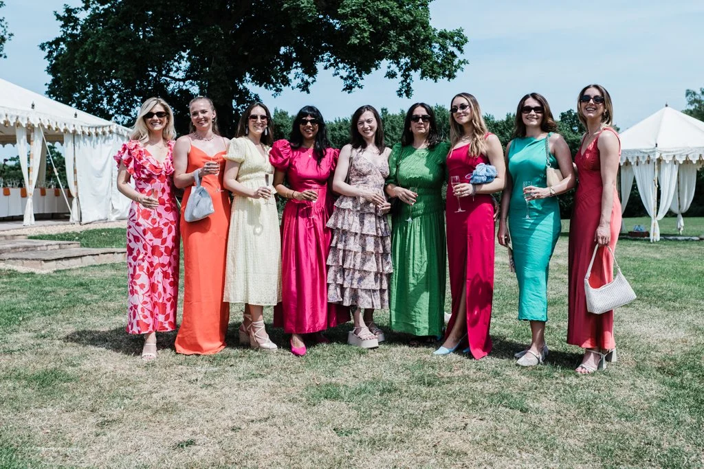 Group of women in colorful summer dresses standing outdoors with white tents and a tree in the background.