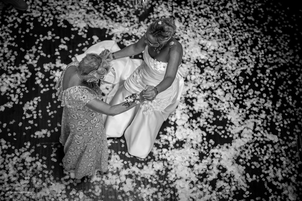 Two women, one in a floral dress and the other in a wedding dress, dancing together on a floor scattered with flower petals.