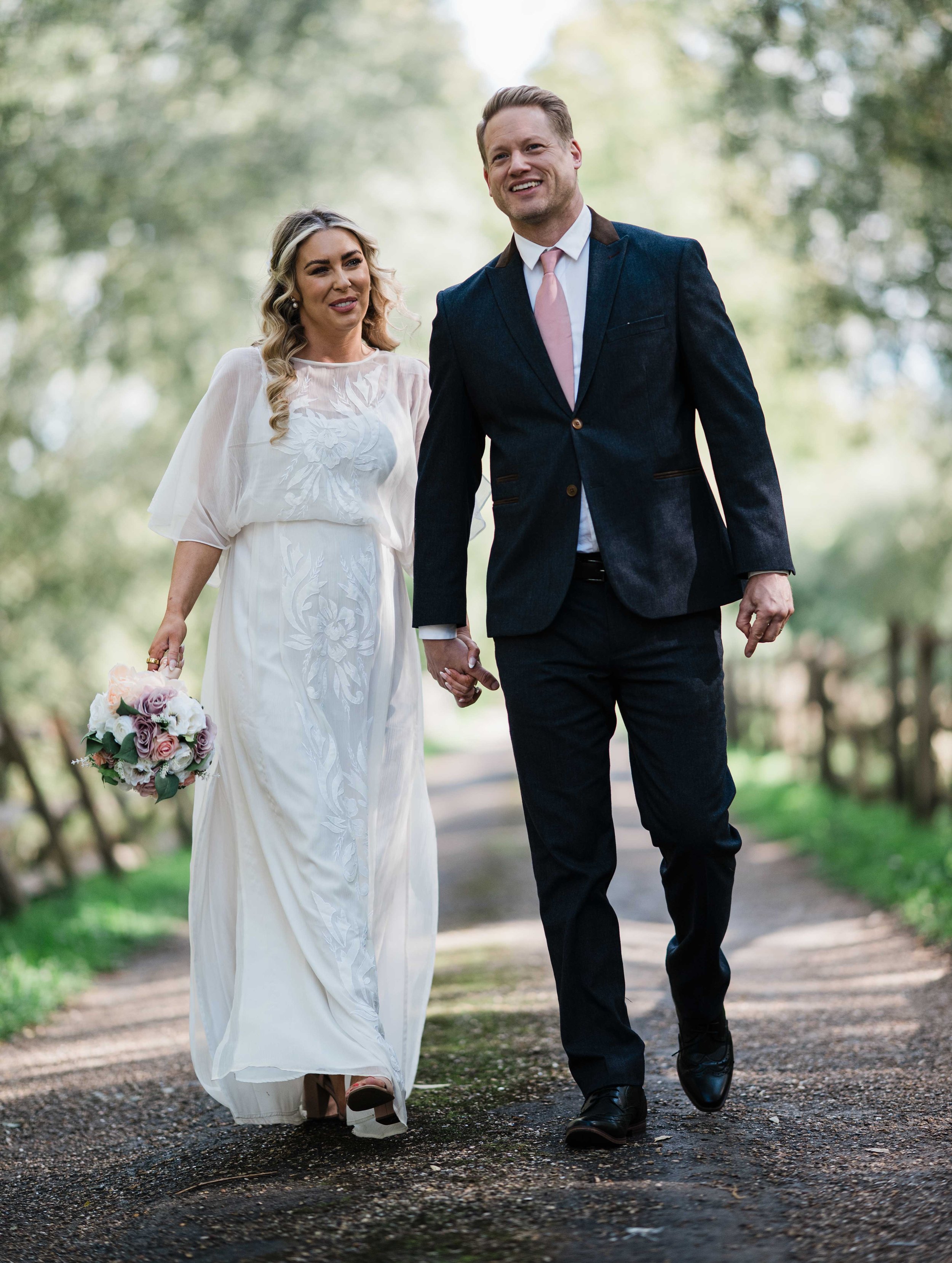 A couple dressed in wedding attire walking hand in hand outdoors along a wooded path, with the woman holding a bouquet of flowers.