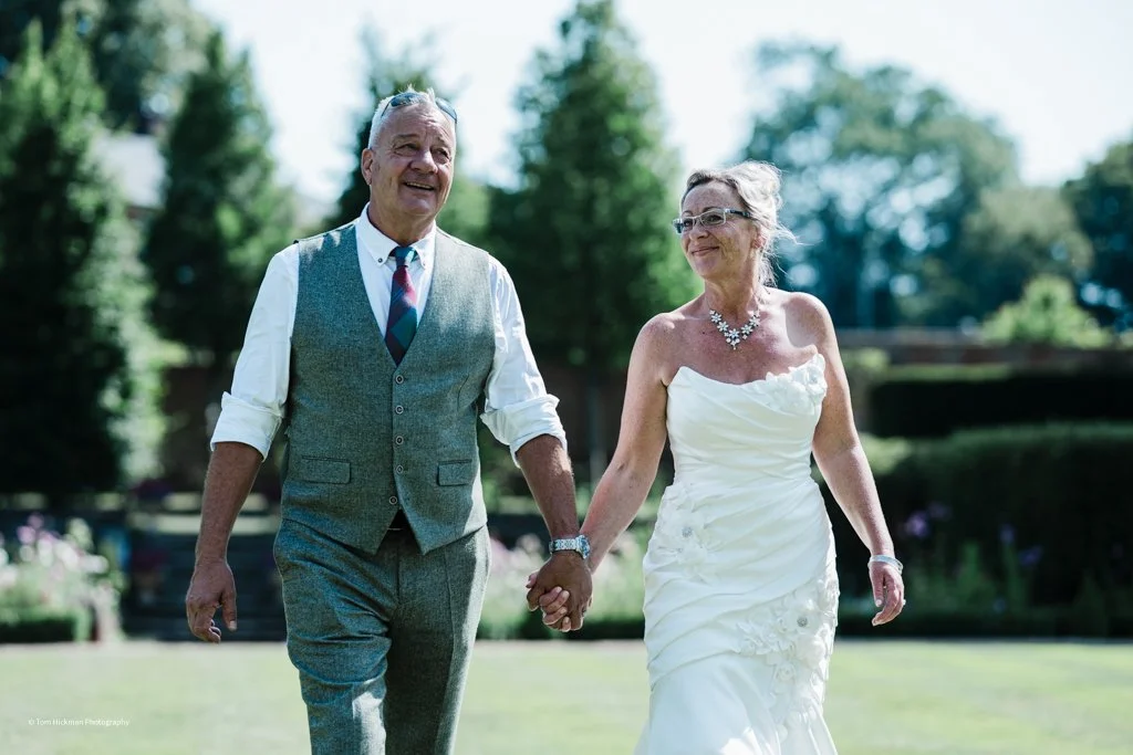 An elderly couple holding hands and smiling while walking outdoors on a sunny day, with lush green trees and grass in the background. The woman is wearing a white wedding dress with jewelry, and the man is dressed in a gray vest and pants with a roll