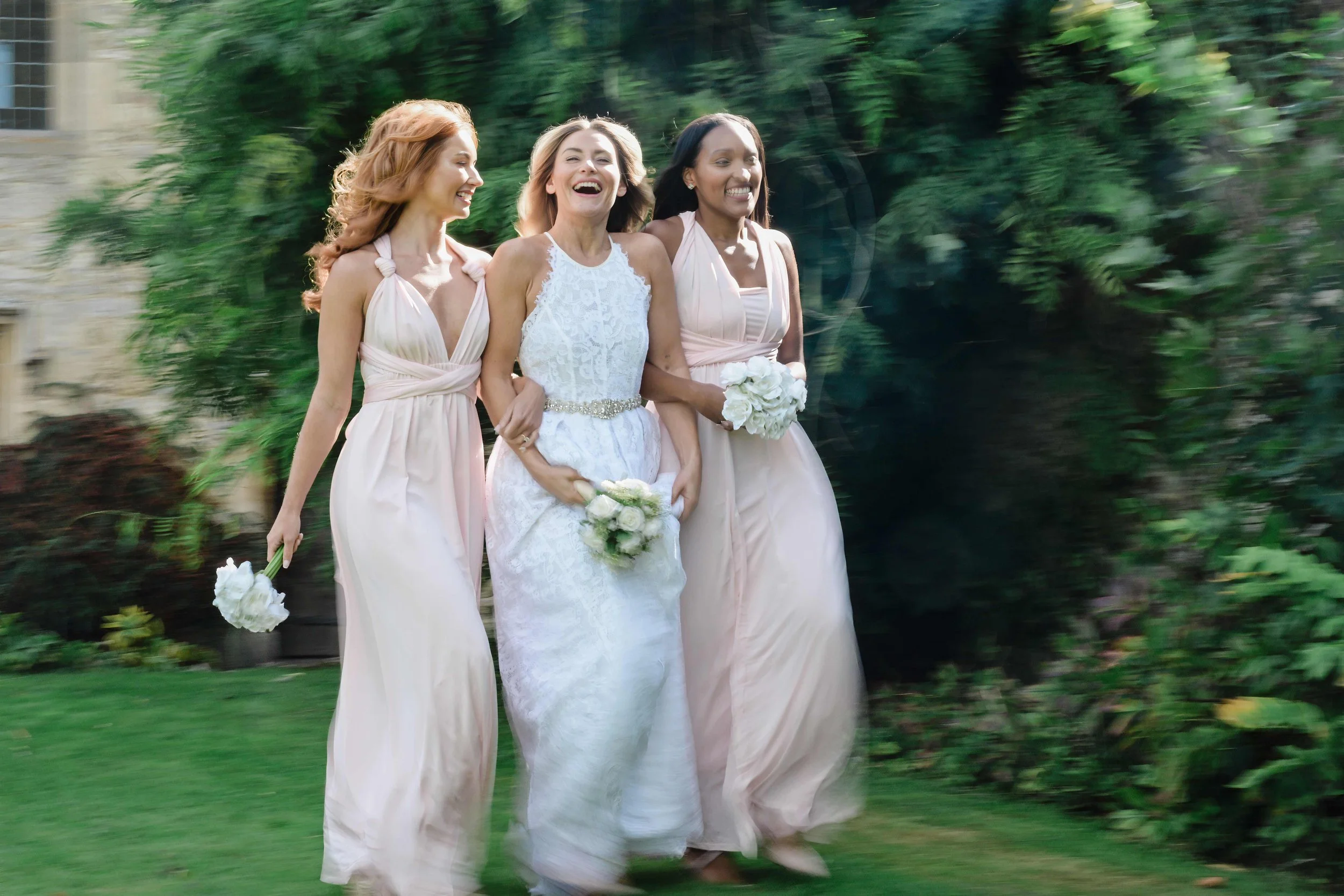 A bride in a white lace wedding dress and two bridesmaids in blush-colored dresses walk outdoors holding bouquets of white flowers, smiling and laughing.