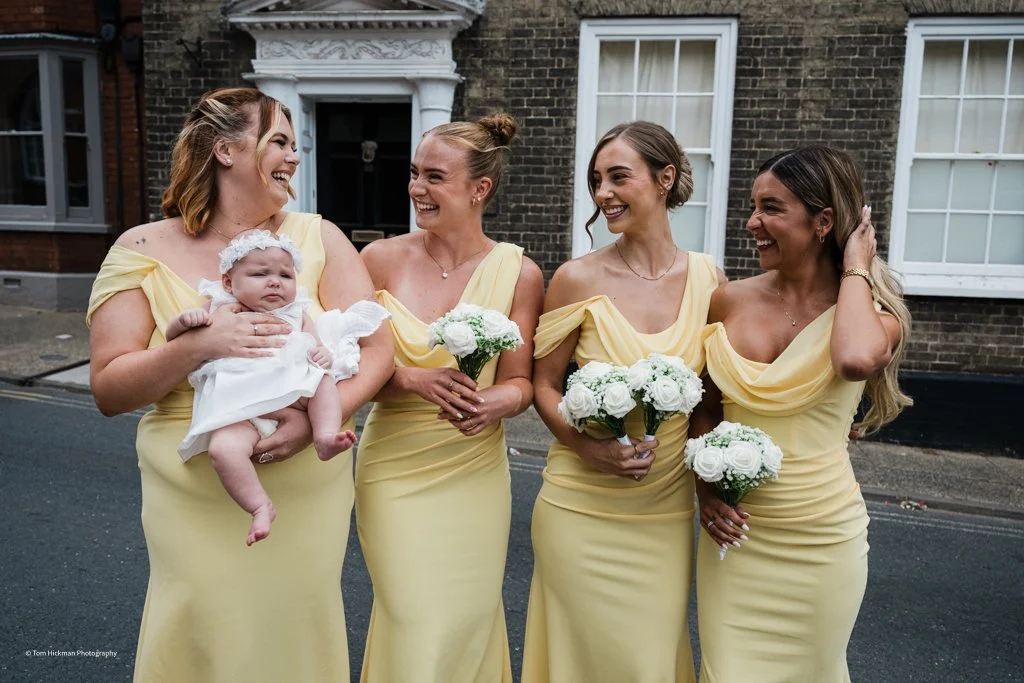 Four women in matching yellow dresses, holding bouquets of white flowers, standing outside a brick building. One woman is holding a baby girl dressed in white. They are smiling and engaging with each other.