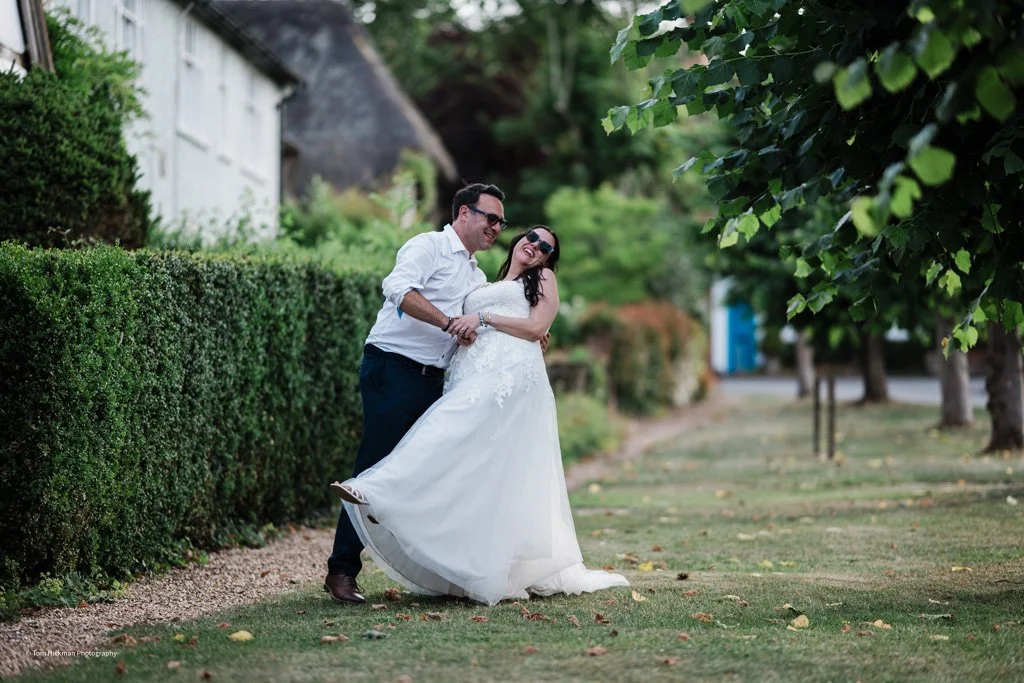 A smiling couple, possibly newlyweds, enjoying a moment outdoors on a grassy path with trees and bushes around. The woman is wearing a white wedding dress and sunglasses, and the man is dressed in a white shirt and dark pants.