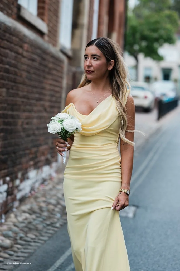 A woman in a yellow strapless dress holding a bouquet of white roses walking along a street with brick buildings.