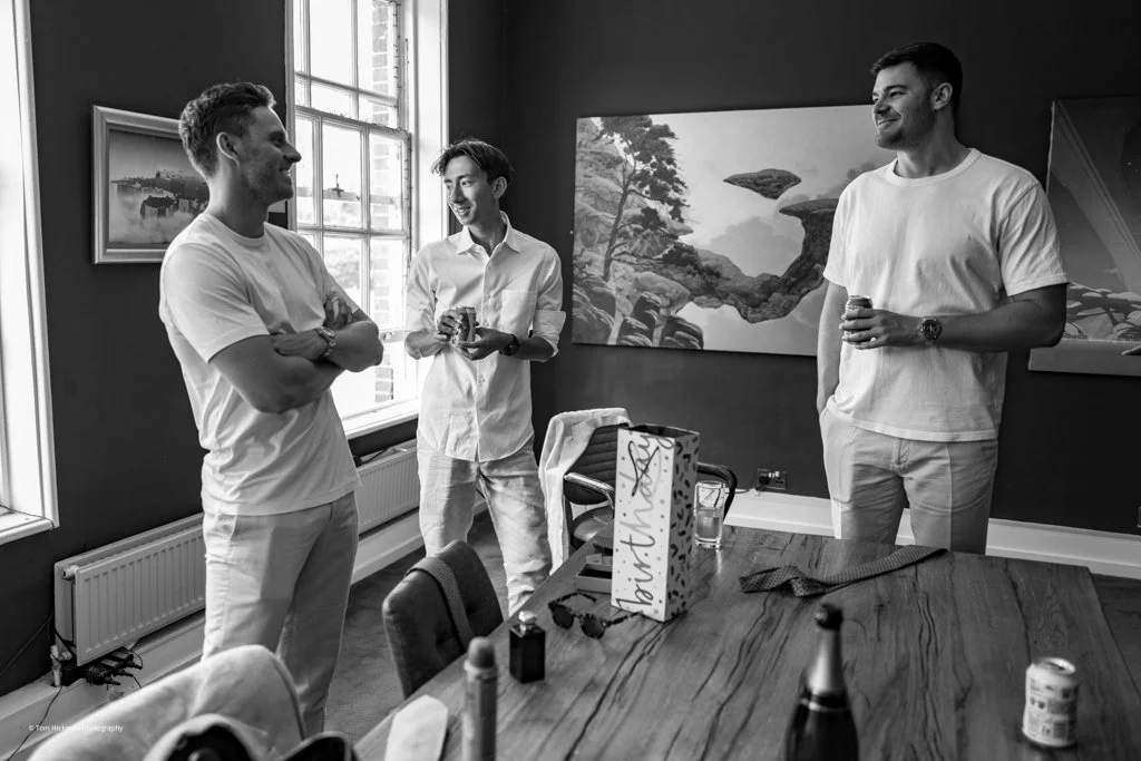 Three men in casual clothes are standing and talking in a room, with two of them smiling at each other. One is holding a drink, and another has his arms crossed. The room has a large window, artwork on the walls, a table with a gift box, glasses, and