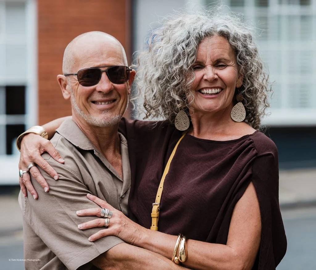 A smiling older man with sunglasses and a woman with curly gray hair, both embracing and standing outdoors in an urban setting.