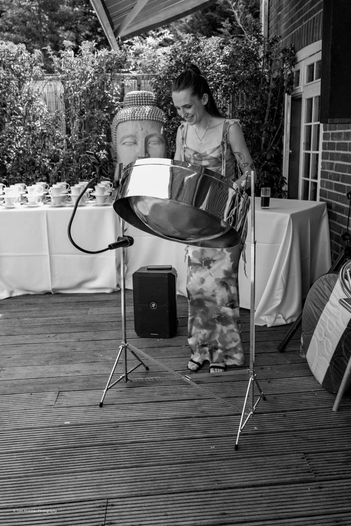 A woman playing a steel pan drum at an outdoor event. She is smiling and standing on a wooden deck with tables in the background. There is a large Buddha head statue and several teacups on the table behind her.