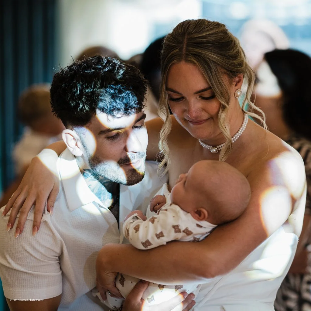 A woman holding a baby and a man with tattoos looking at the baby, with a background of people at an event.