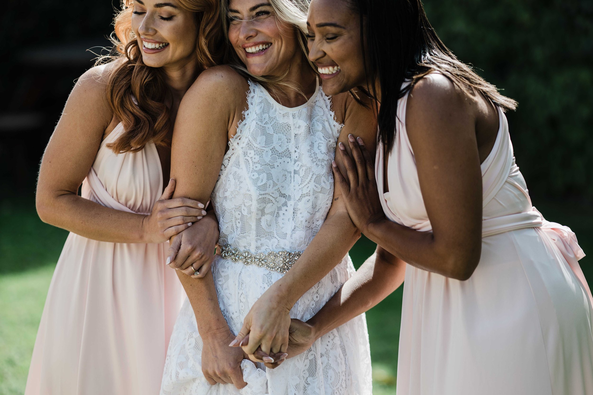 Four women smiling and embracing each other outdoors, with one in a white lace dress and the others in light pink dresses.