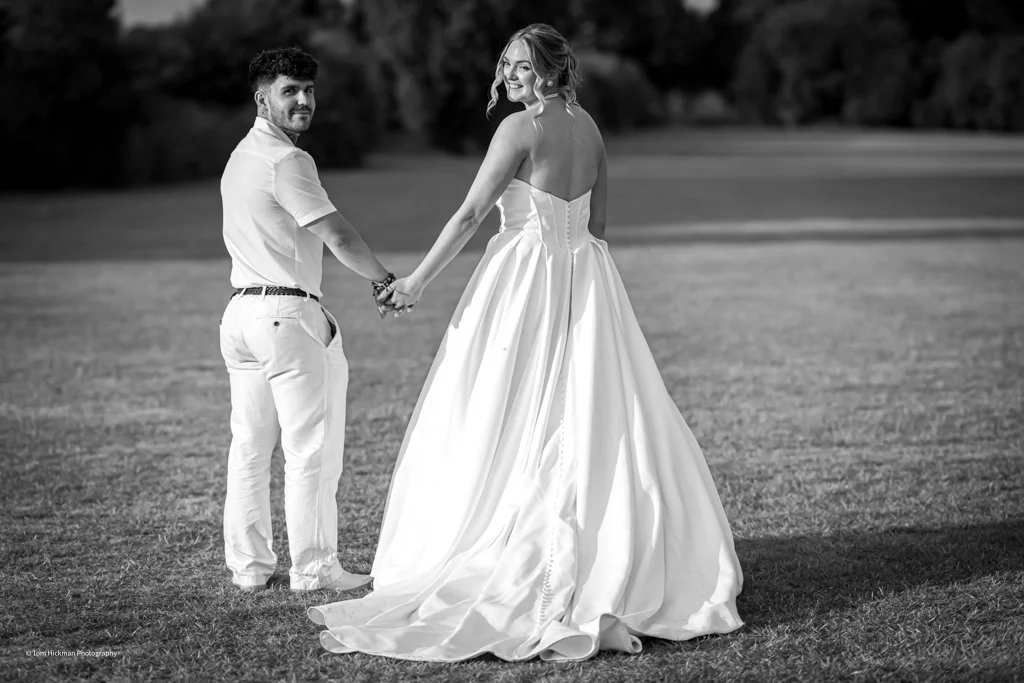 A couple holding hands, the woman in a wedding dress and the man in white pants and a shirt, standing on a grassy field outdoors.