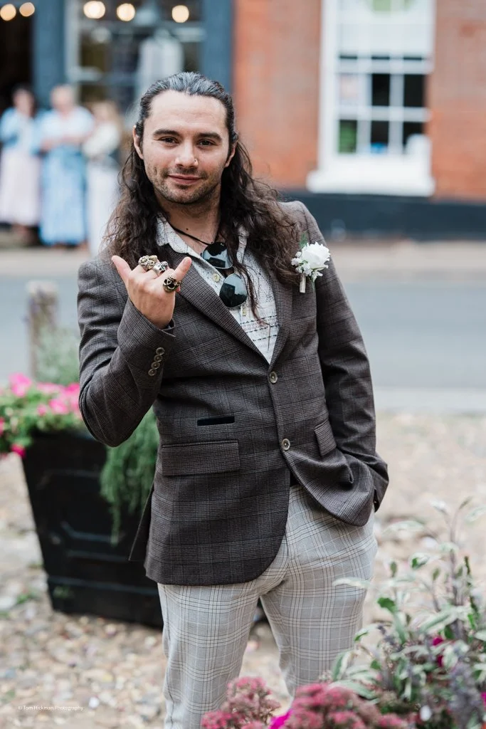 Man with long hair wearing a suit with rings, a boutonniere, and sunglasses, standing outdoors on a street with a woman in the background.