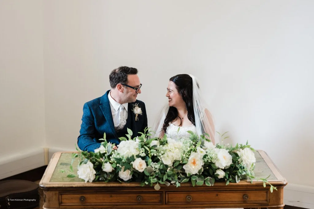 A bride and groom sitting at a wooden table with a floral arrangement, smiling and looking at each other.