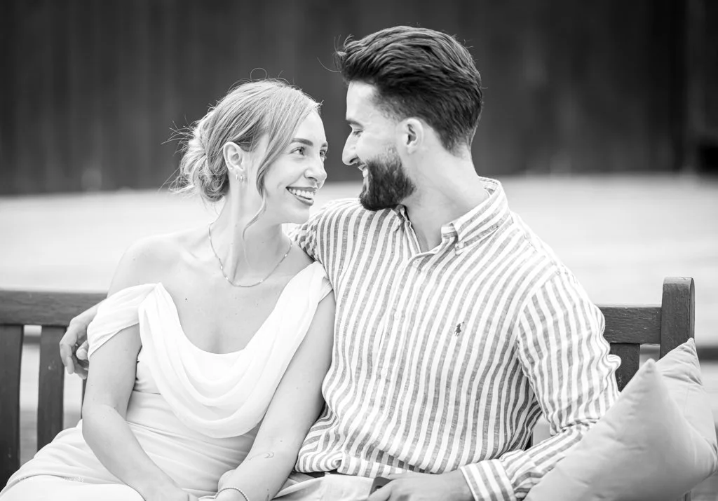 A couple sitting closely on a wooden bench, facing and looking at each other, smiling, in an outdoor setting.
