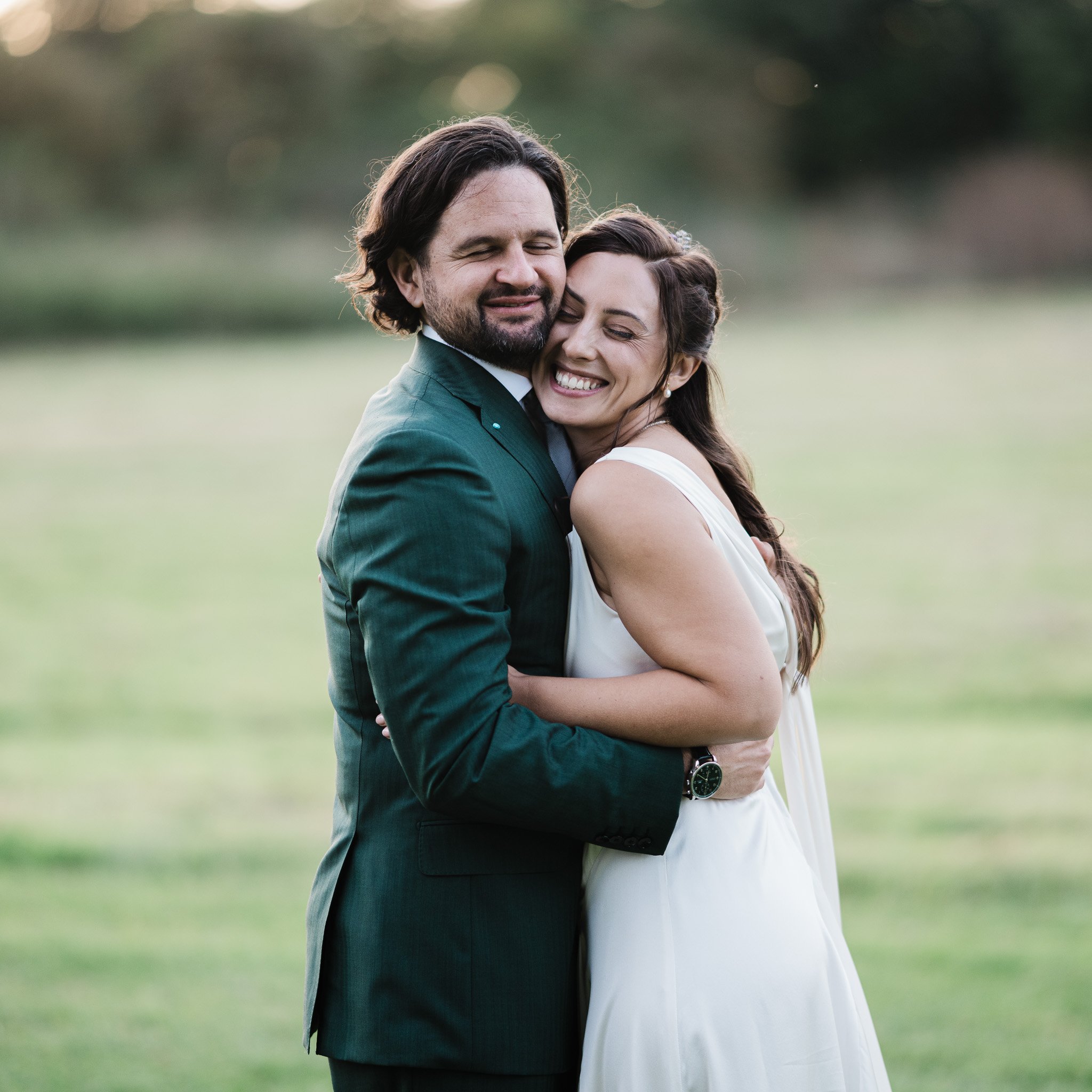 A man and woman embracing outdoors with a blurred green background, both smiling and with eyes closed.