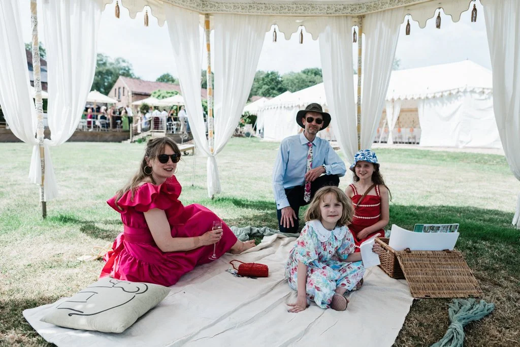 A family enjoying a picnic under a white canopy tent at an outdoor event, with women and children sitting on a blanket on the grass.