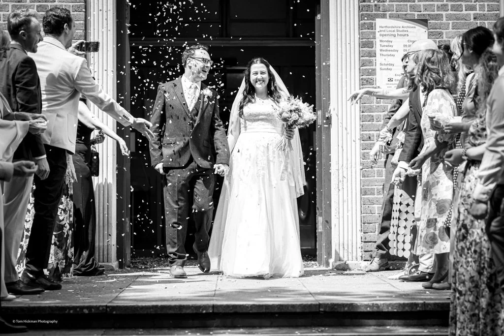 Bride and groom exiting a building, smiling, surrounded by guests throwing confetti in celebration.