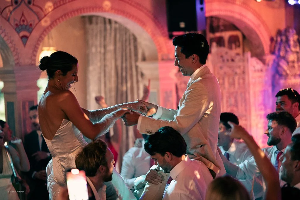 A bride and groom share a joyful dance during their wedding reception, surrounded by guests in a decorated venue with colorful lighting.