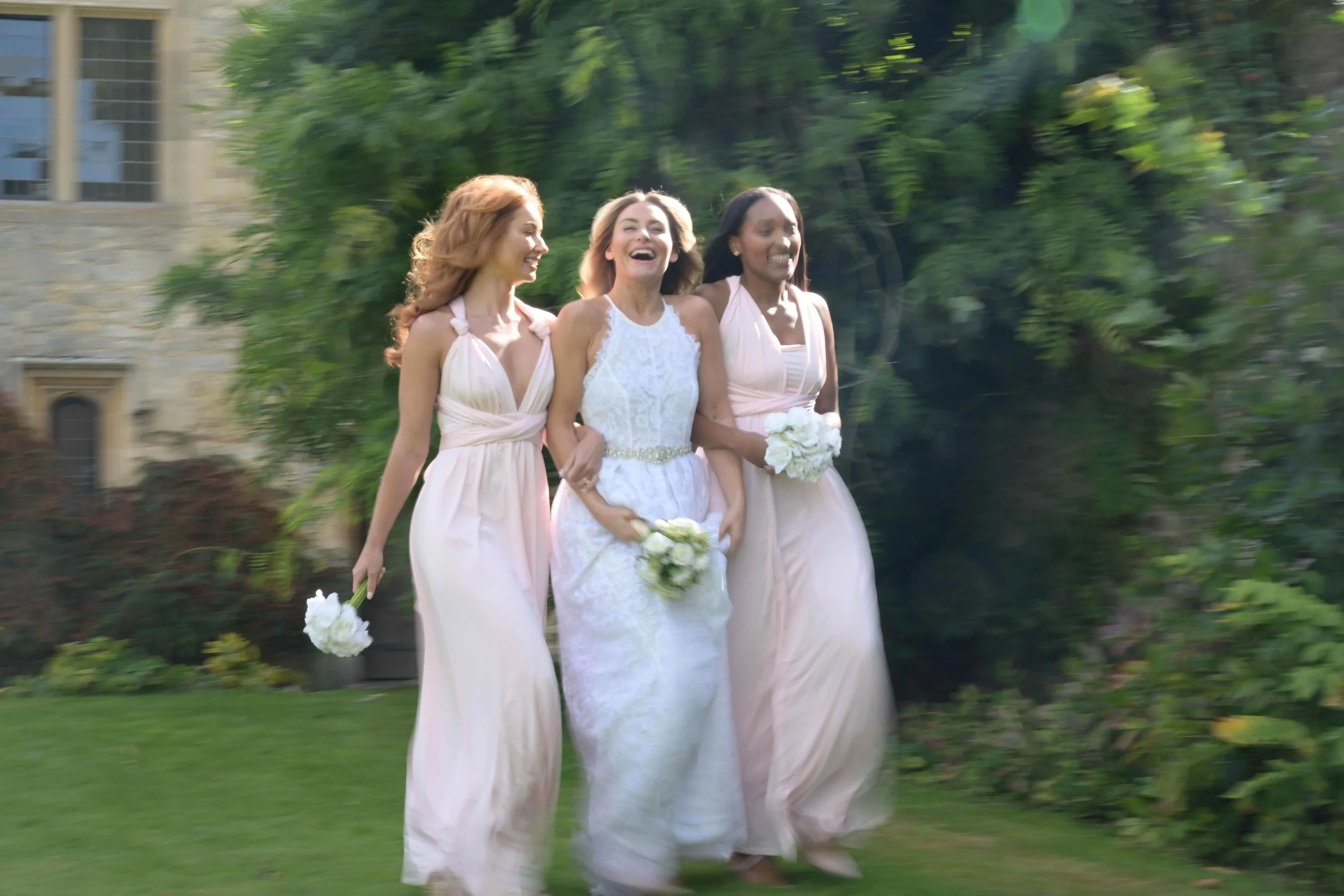 Three women, one in a white wedding dress and two in pastel pink bridesmaid dresses, are smiling and walking outdoors in a garden setting, with greenery and a stone building in the background.
