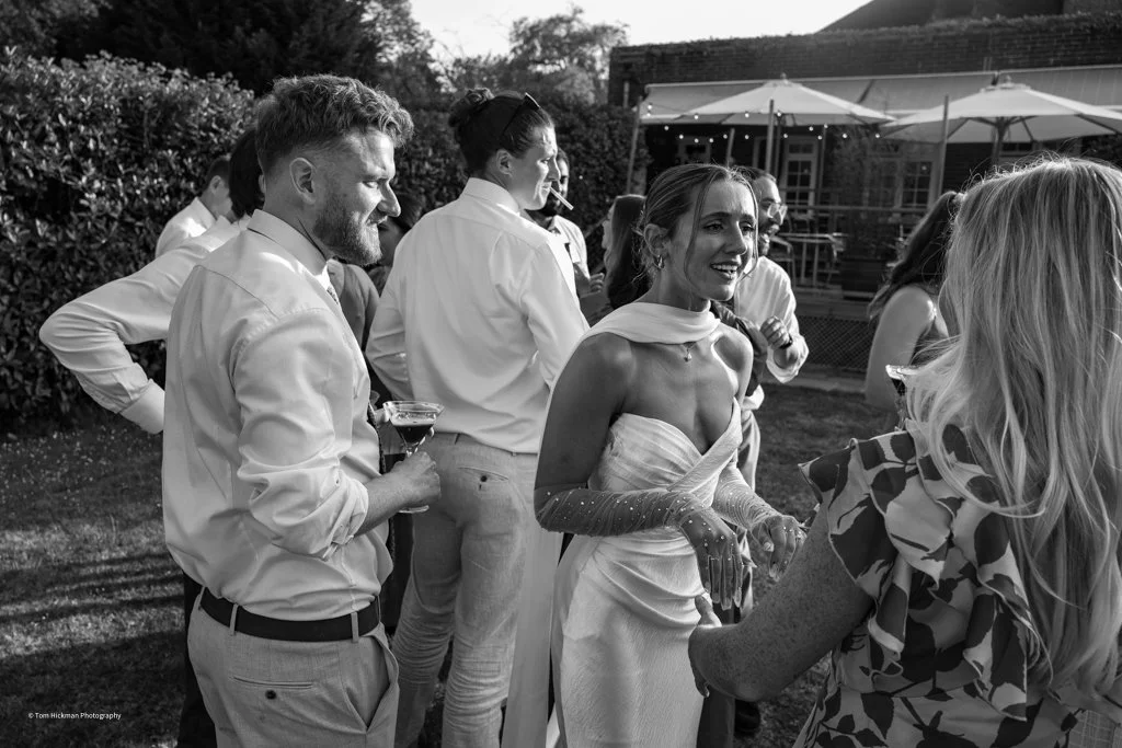Guests at a wedding reception outdoors, including a woman in a white dress and a man in a shirt holding a drink, engaging in conversation.