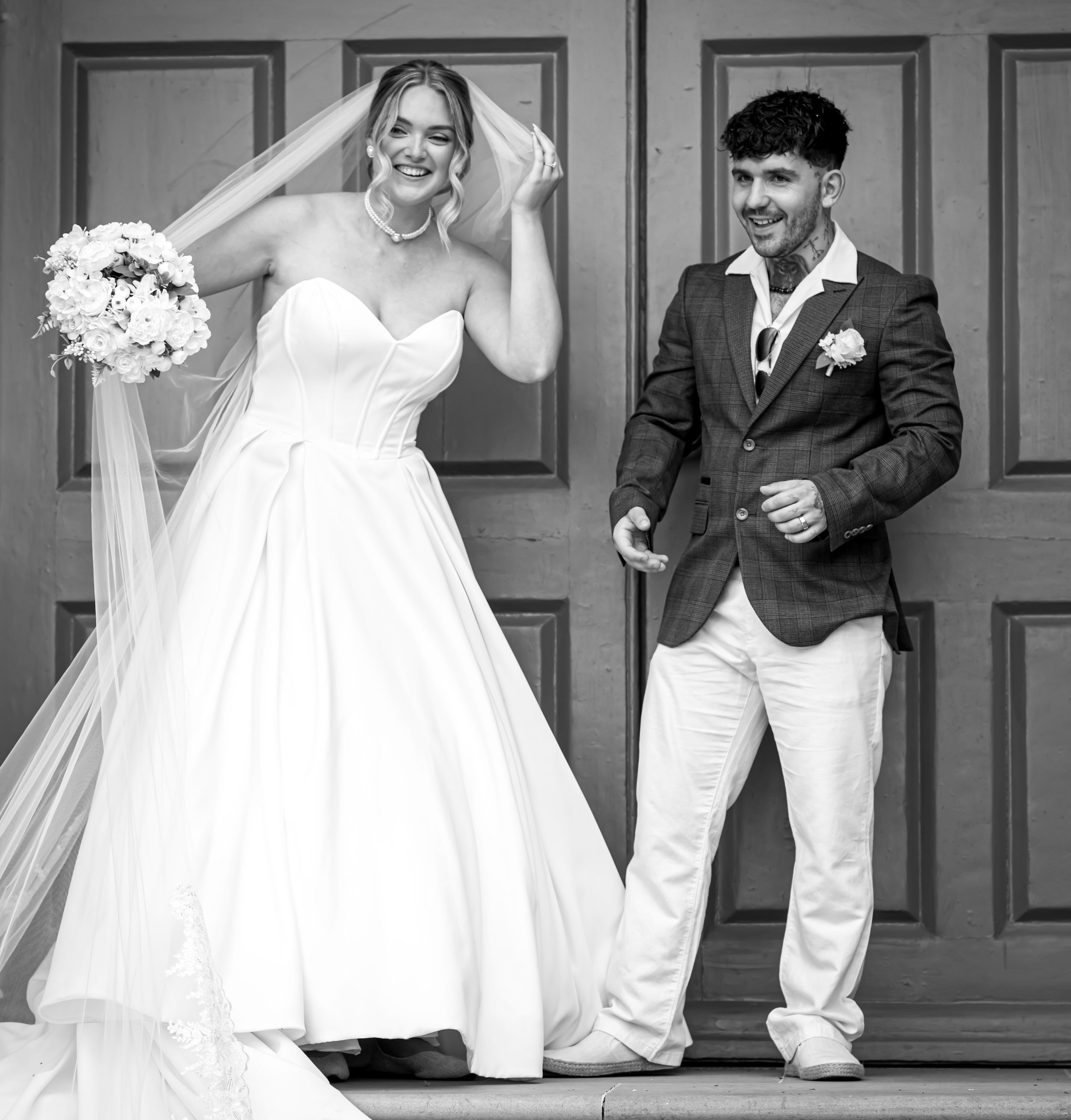 Black-and-white photo of a bride and groom standing in front of a wooden door, both smiling and dressed in wedding attire, with the bride holding a bouquet of flowers.
