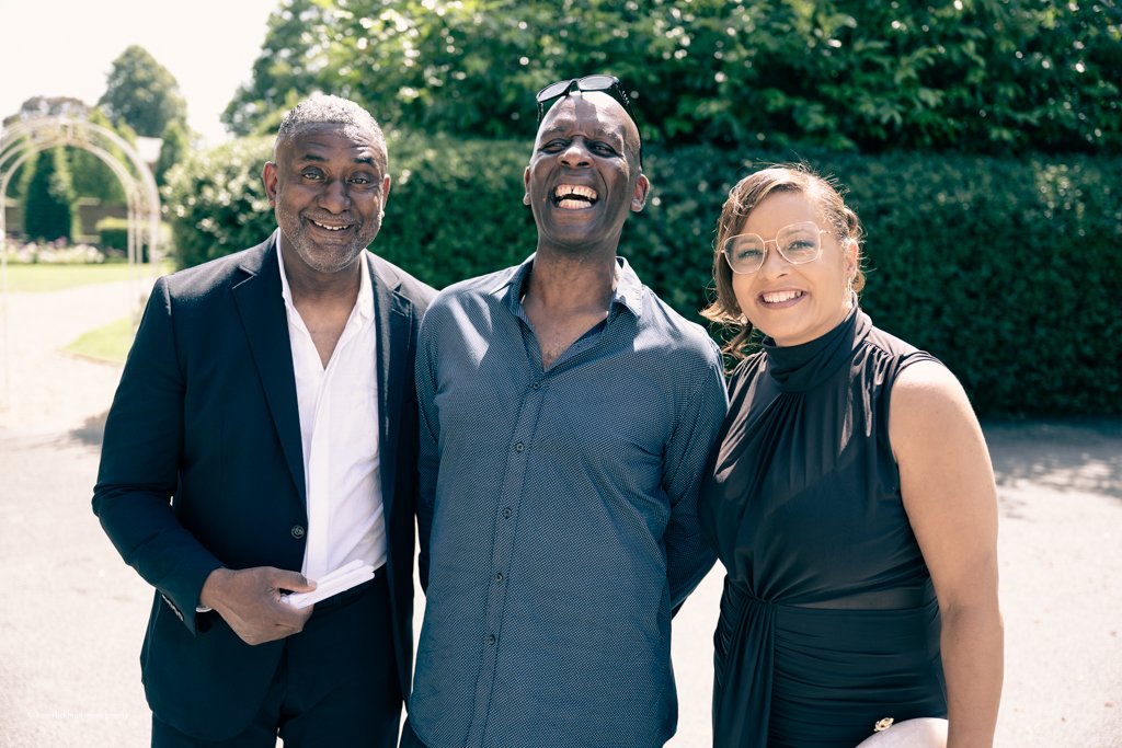 Three people standing outdoors on a sunny day, smiling and enjoying each other's company, with greenery and a park structure in the background.
