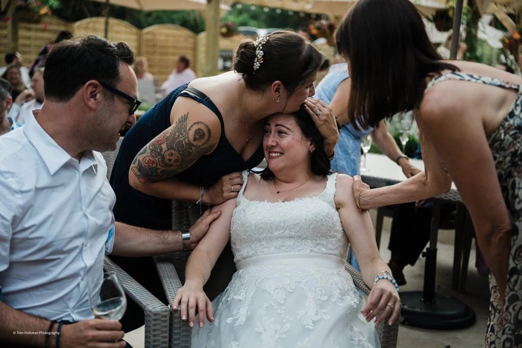 A bride in a white lace dress sitting outdoors, smiling as a woman kisses her on the forehead and others gather around in celebration.