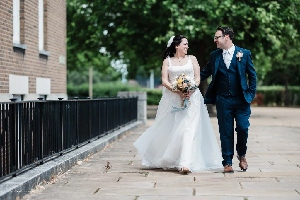 A bride and groom walking outdoors on a paved pathway, smiling at each other, with green trees in the background. The bride is holding a bouquet and wearing a white wedding dress, and the groom is in a blue suit with brown shoes.
