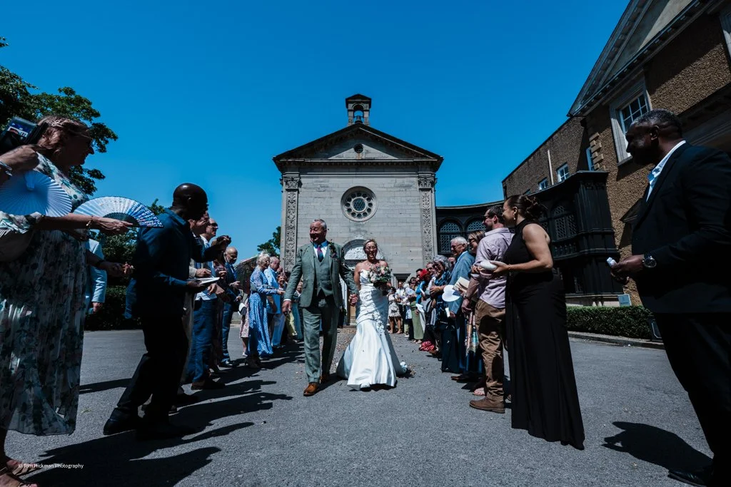 A wedding procession outside a church with guests lining both sides, the bride walking with her father, all under a clear blue sky.